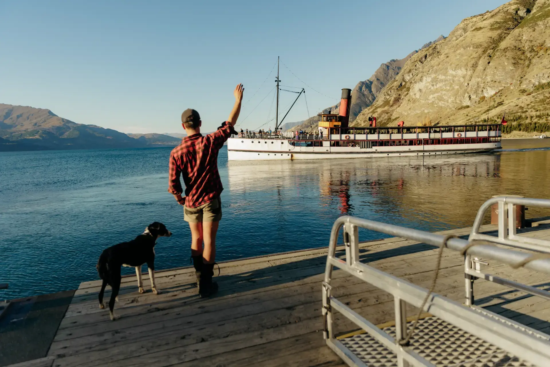 A man in a plaid shirt, with a border collie standing at his feet, waves from the shorw to a historic steamship sailing away on a lake on a sunny day. 