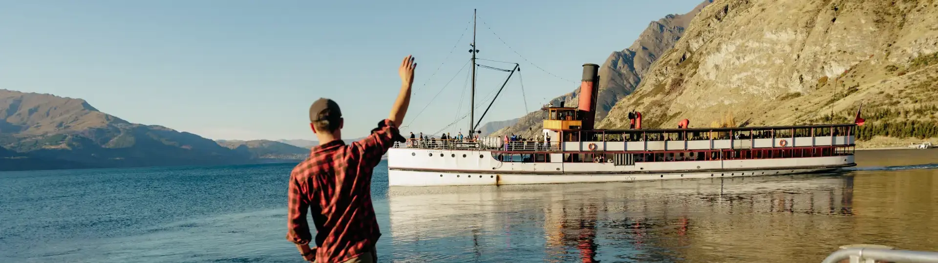 A man in a plaid shirt, with a border collie standing at his feet, waves from the shorw to a historic steamship sailing away on a lake on a sunny day. 