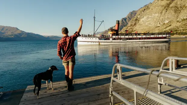 A man in a plaid shirt, with a border collie standing at his feet, waves from the shorw to a historic steamship sailing away on a lake on a sunny day. 