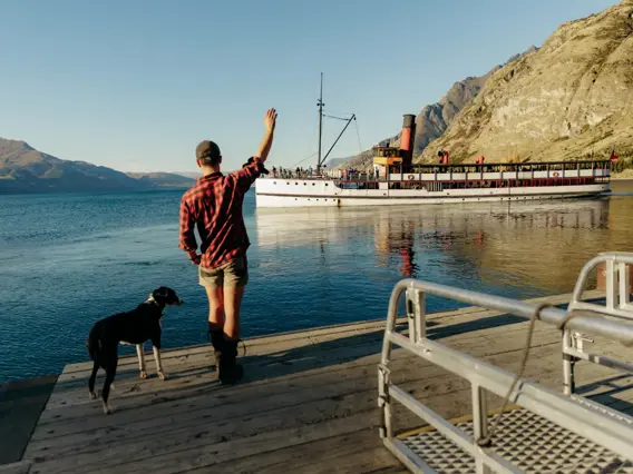 A man in a plaid shirt, with a border collie standing at his feet, waves from the shorw to a historic steamship sailing away on a lake on a sunny day. 