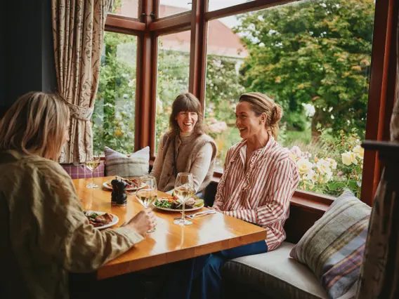 Three women enjoying a meal and wine beside a garden window.
