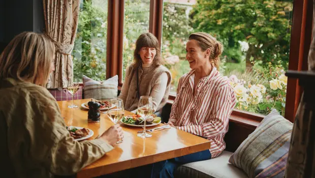 Three women enjoying a meal and wine beside a garden window.