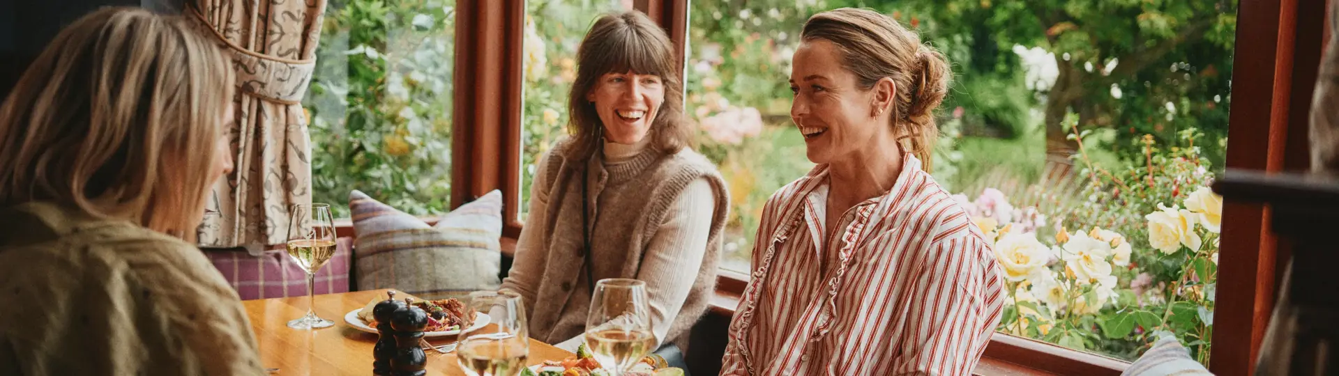 Three women enjoying a meal and wine beside a garden window.