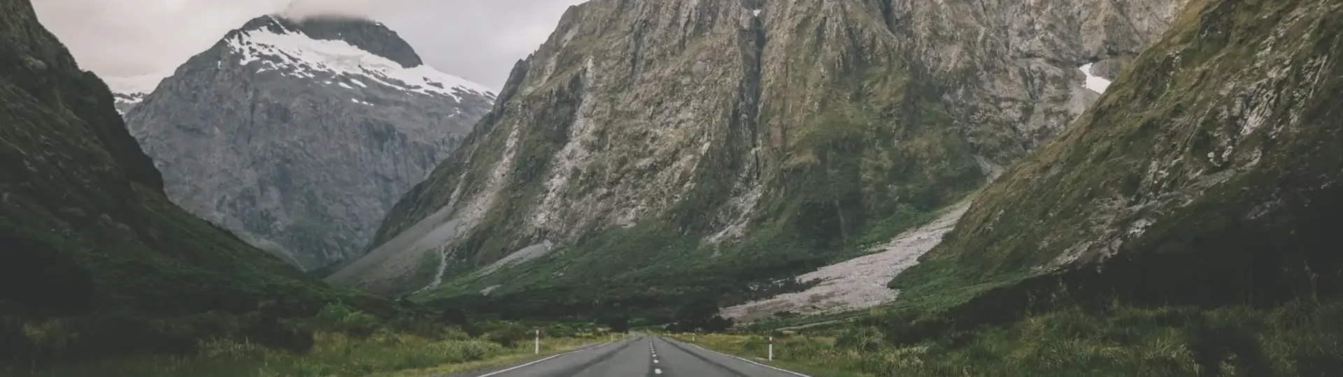 A straight two-lane road cutting through a dramatic alpine valley toward jagged, snow-dusted peaks.