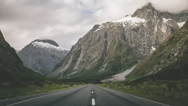 A straight two-lane road cutting through a dramatic alpine valley toward jagged, snow-dusted peaks.