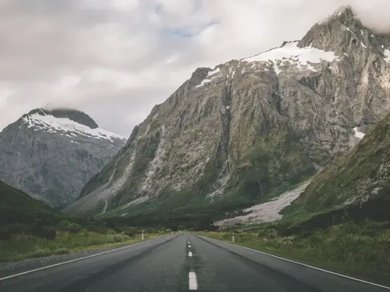 A straight two-lane road cutting through a dramatic alpine valley toward jagged, snow-dusted peaks.