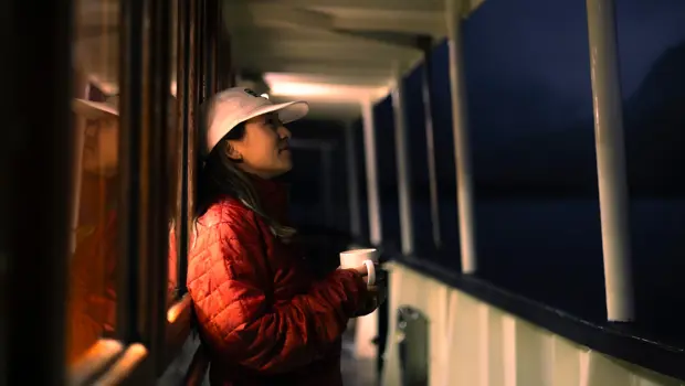 Woman holding a mug on the deck of a boat at dusk.