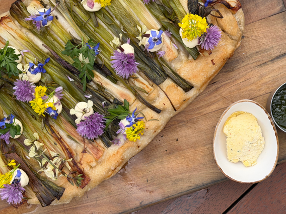 Spring leek focaccia topped with herbs and edible flowers, served with butter and green herb sauce on a wooden board.