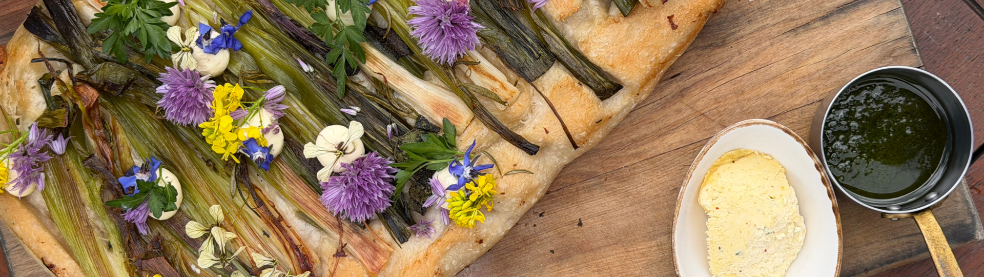 Spring leek focaccia topped with herbs and edible flowers, served with butter and green herb sauce on a wooden board.