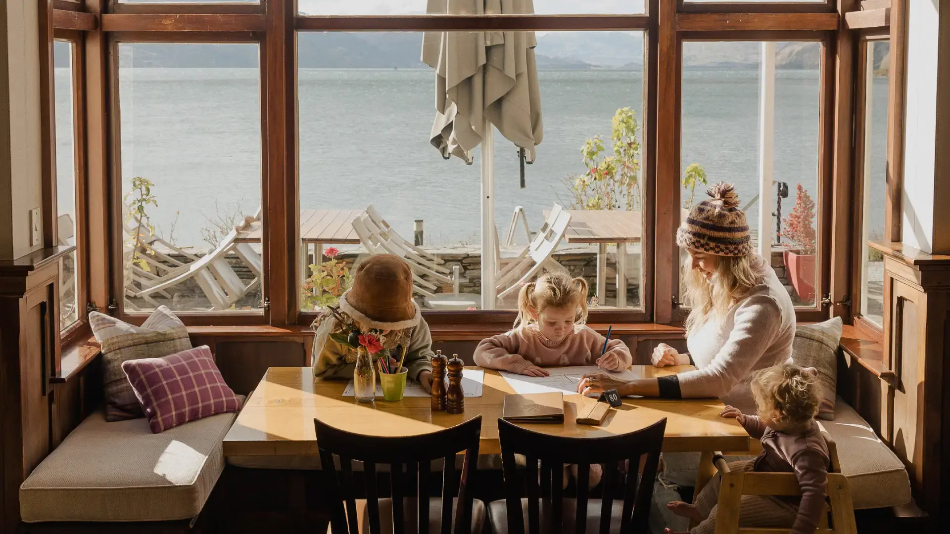 Mother and three young children sit at a sunny table inside Walter Peak Homestead, drawing and relaxing by large windows overlooking the lake.
