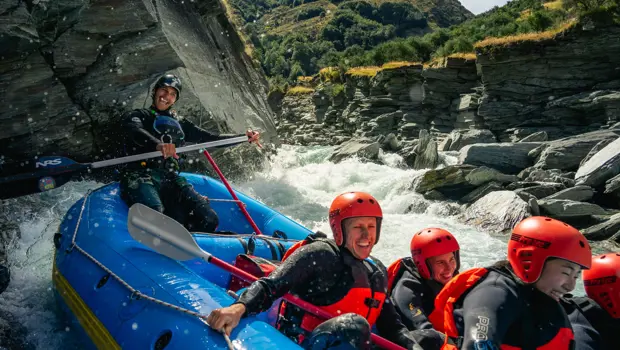 A group of smiling rafters in red helmets and wetsuits navigating white water rapids through a rocky gorge near Queenstown.