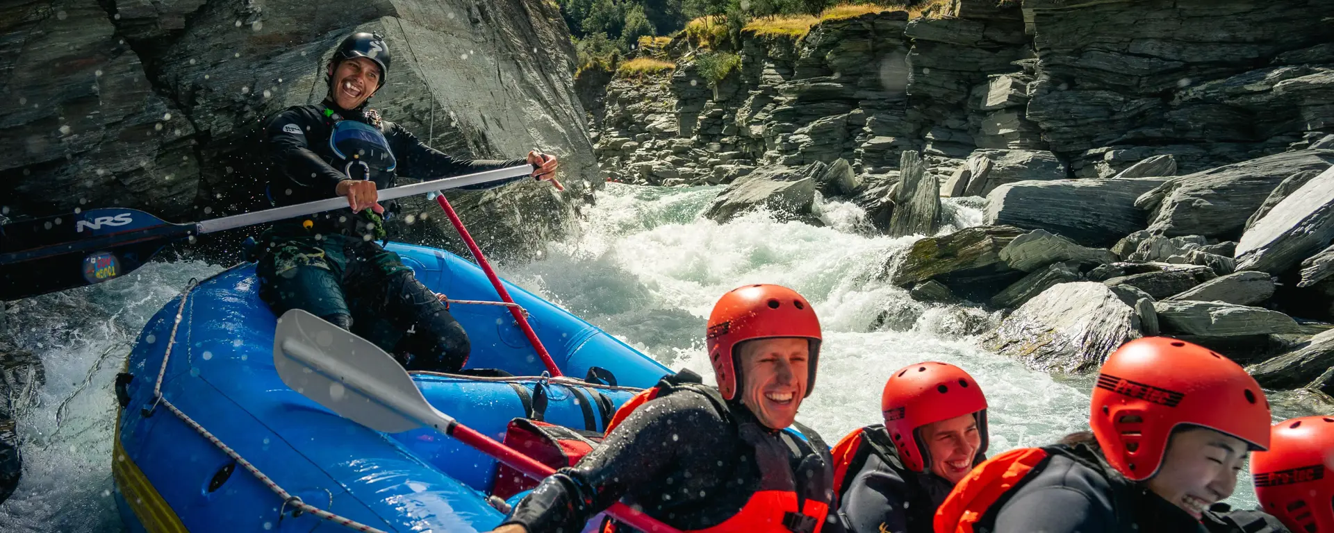 A group of smiling rafters in red helmets and wetsuits navigating white water rapids through a rocky gorge near Queenstown.