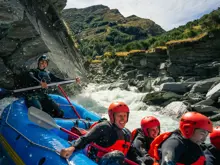 A group of smiling rafters in red helmets and wetsuits navigating white water rapids through a rocky gorge near Queenstown.