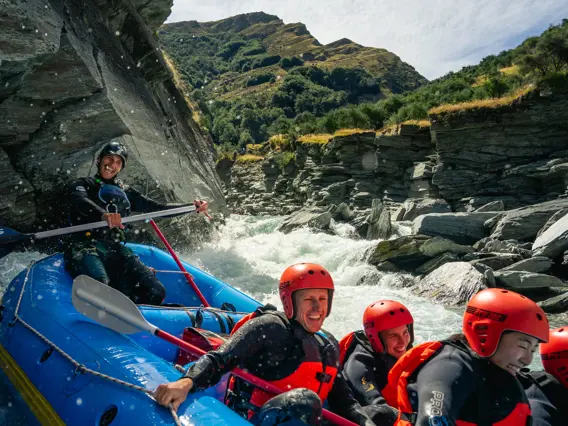 A group of smiling rafters in red helmets and wetsuits navigating white water rapids through a rocky gorge near Queenstown.