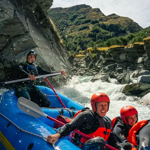 A group of smiling rafters in red helmets and wetsuits navigating white water rapids through a rocky gorge near Queenstown.