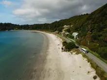 A small white tour bus drives round a coastal road next to a sandy golden beach and blue water.
