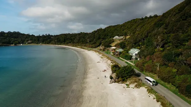 A small white tour bus drives round a coastal road next to a sandy golden beach and blue water.