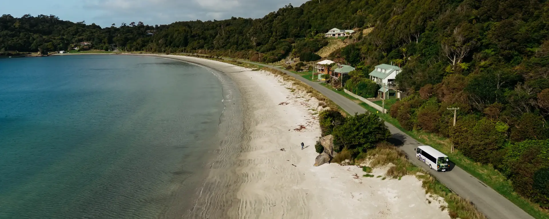 A small white tour bus drives round a coastal road next to a sandy golden beach and blue water.