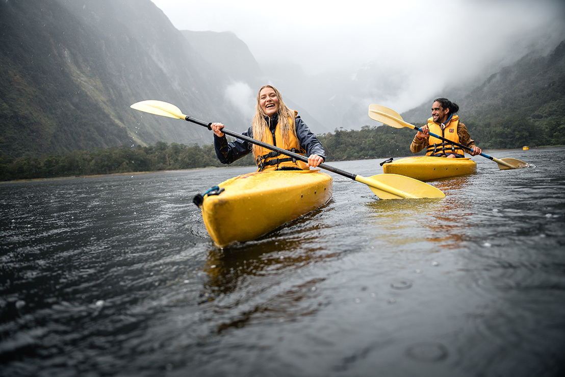 A couple kayak on bright yellow kayaks through a moody Milford Sound 