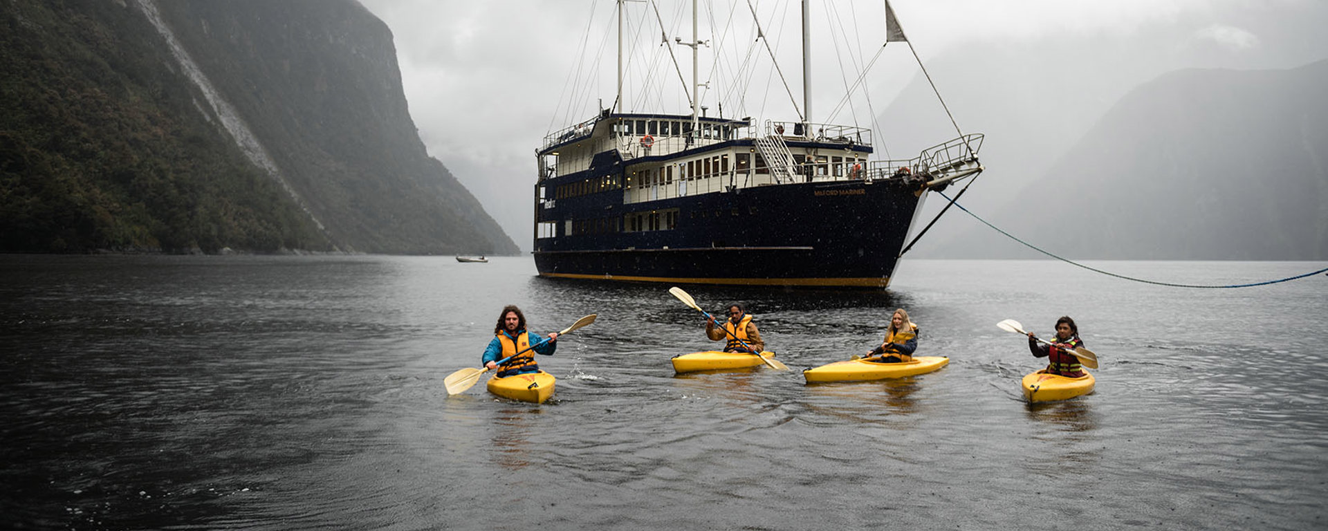 A group of four kayakers floating on Milford Sound in front of the Milford Wanderer boat. 