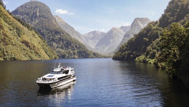 A cruise boat journeys through the water at Doubtful Sound on a clear day, surrounded by mountains 
