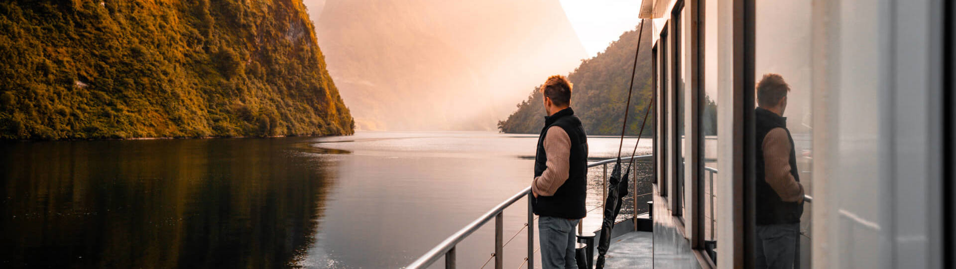 A man stands on the side deck of a boat cruising through Doubtful  Sound at golden hour