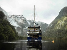 A kayaker paddles next the Fiordland Navigator boat on Doubtful Sound, surrounded by snowy mountains
