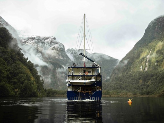 A kayaker paddles next the Fiordland Navigator boat on Doubtful Sound, surrounded by snowy mountains