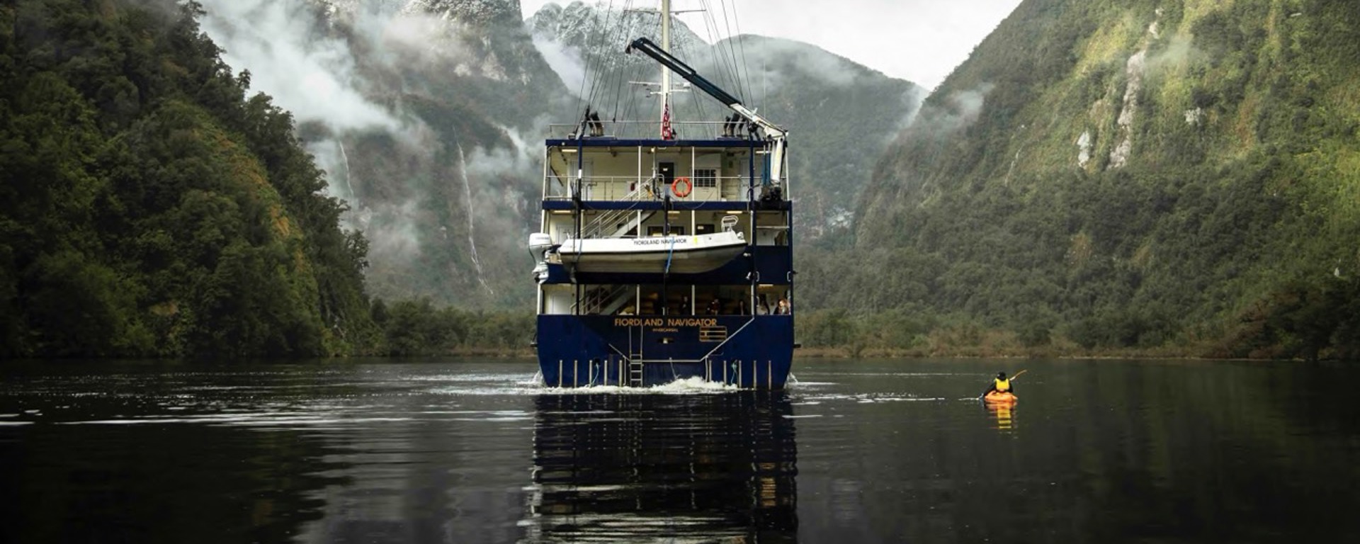 A kayaker paddles next the Fiordland Navigator boat on Doubtful Sound, surrounded by snowy mountains