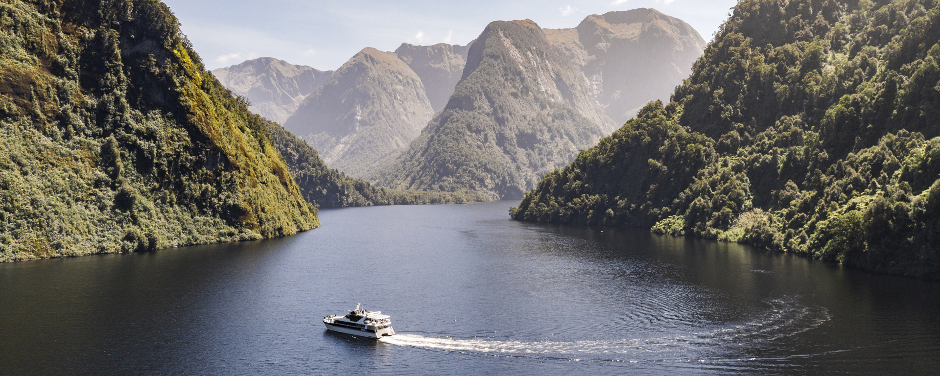 Wide shot of a motorised boat cruising through Doubtful Sound on a bluebird day