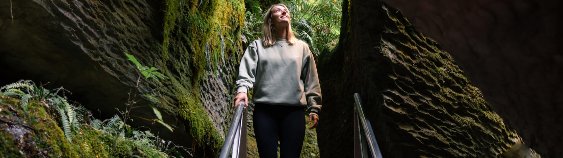 A woman stands on a walkway surrounded by moss-covered rock walls, gazing upward as she enters the lush and enchanting entrance to the Te Anau Glowworm Caves.