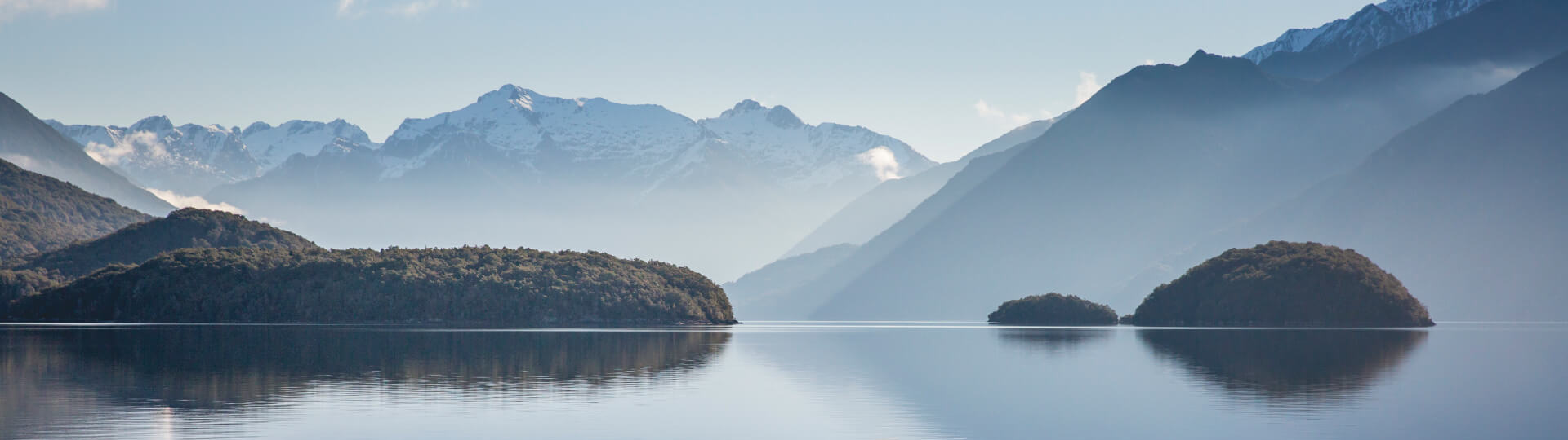  A tranquil lake reflects forested islands and snow-capped mountains under a clear blue sky, creating a stunning backdrop on the start of a Te Anau glowworm caves tour.