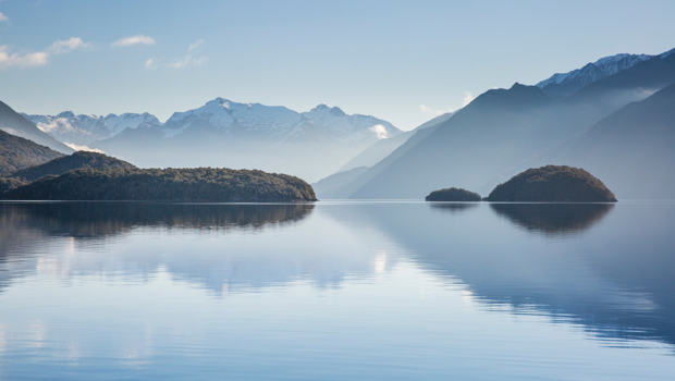  A tranquil lake reflects forested islands and snow-capped mountains under a clear blue sky, creating a stunning backdrop on the start of a Te Anau glowworm caves tour.