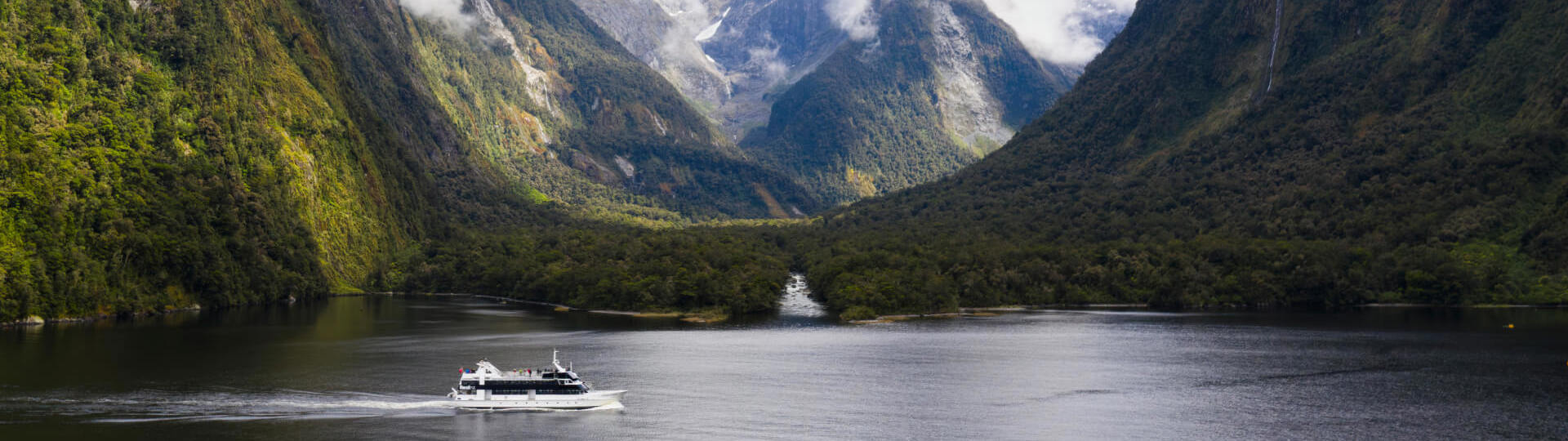 Wide shot of a boat cruising in Milford Sound with snow covered mountains in the background