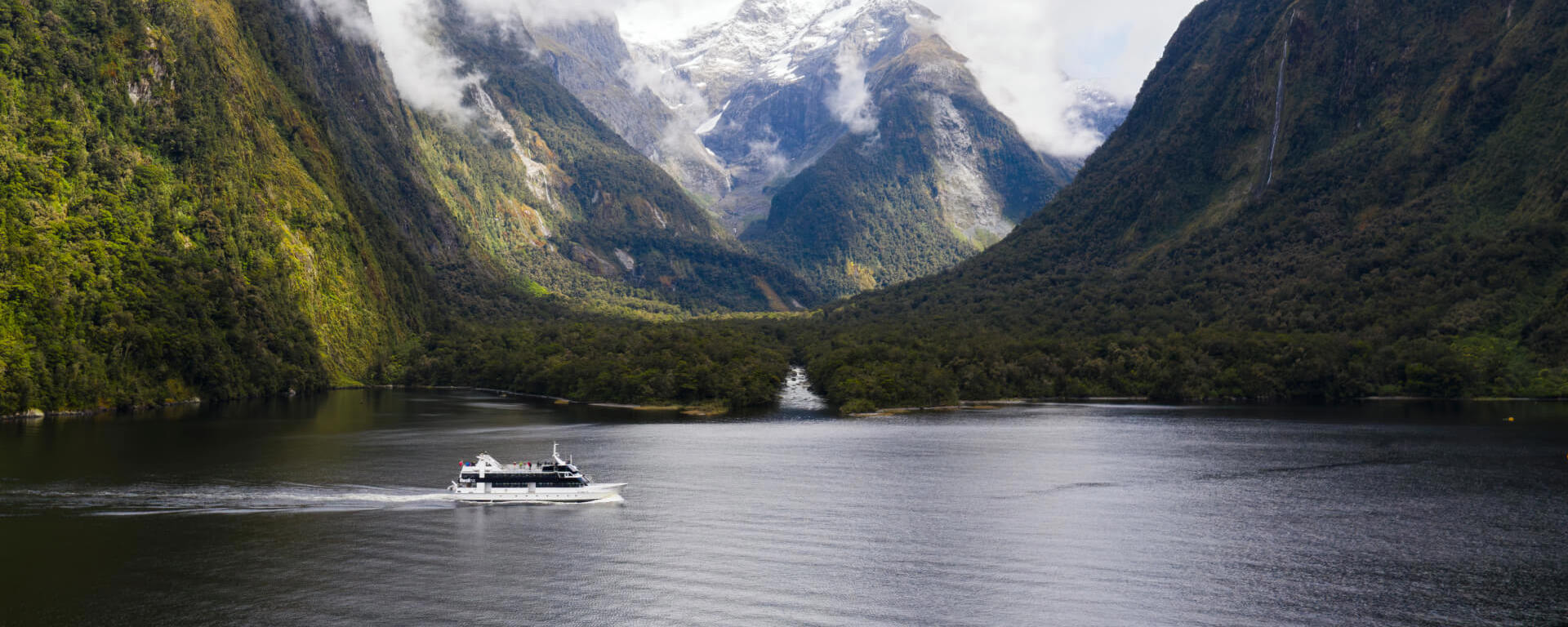 Wide shot of a boat cruising in Milford Sound with snow covered mountains in the background