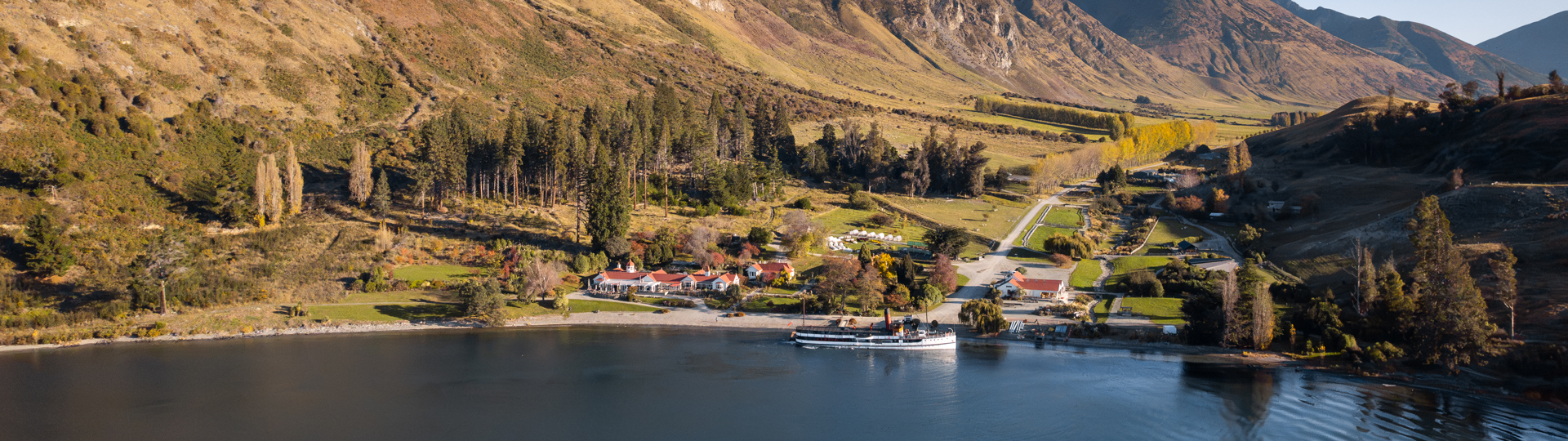 Areal veiw of a lakeside farm surrounded by towering mountains, with a vintage steamship docked in the water. 