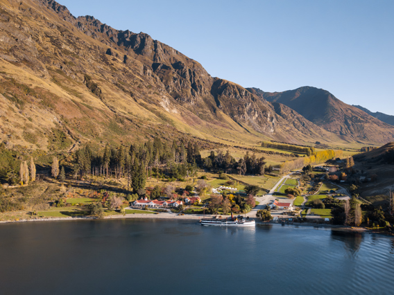 Areal veiw of a lakeside farm surrounded by towering mountains, with a vintage steamship docked in the water. 
