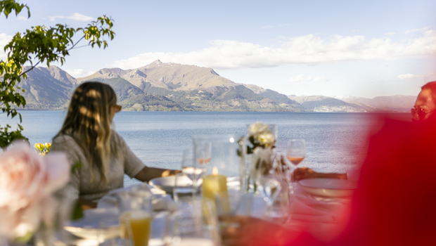 A woman sitting at an outdoor table looks out over Lake Whakatipu at Queenstown in the distance. 