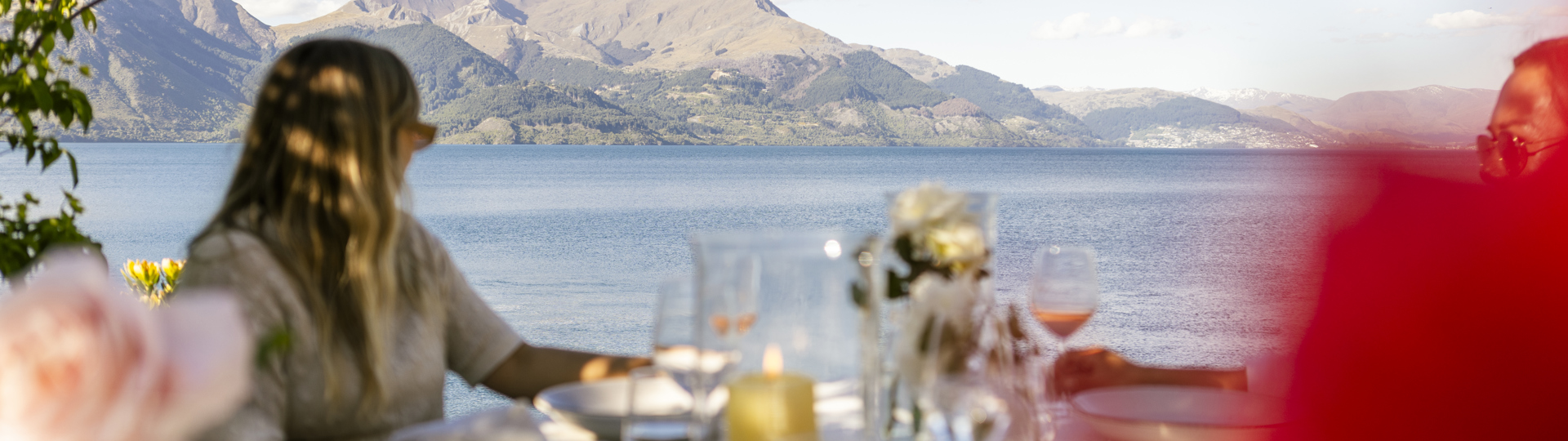 A woman sitting at an outdoor table looks out over Lake Whakatipu at Queenstown in the distance. 