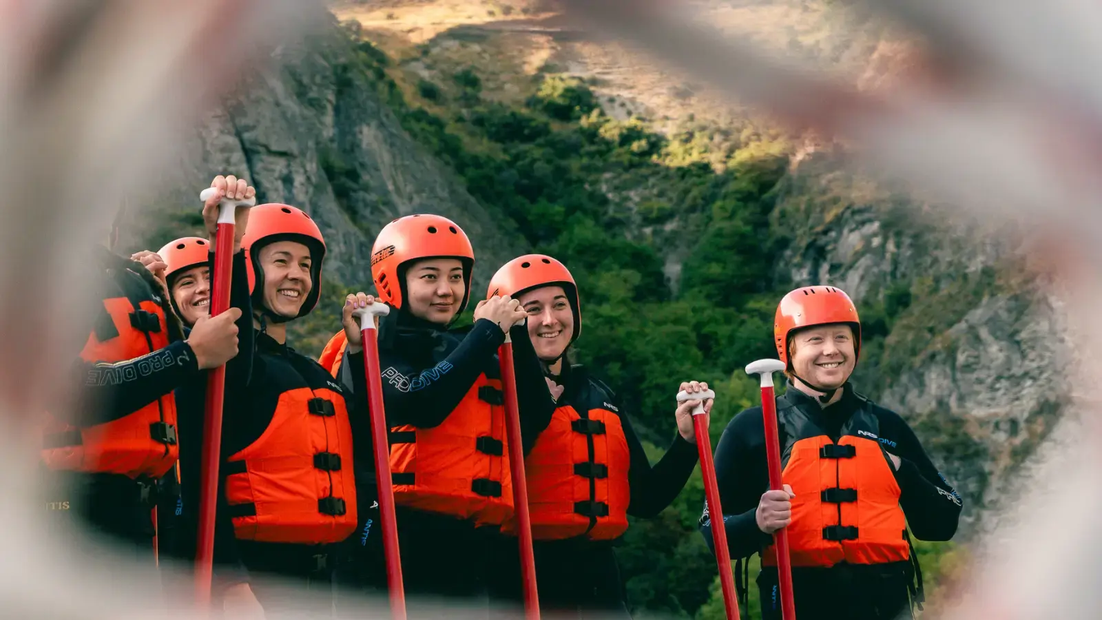 A group of rafters stand wearing orange life jackets, helmets, wesuits and holding paddles. 