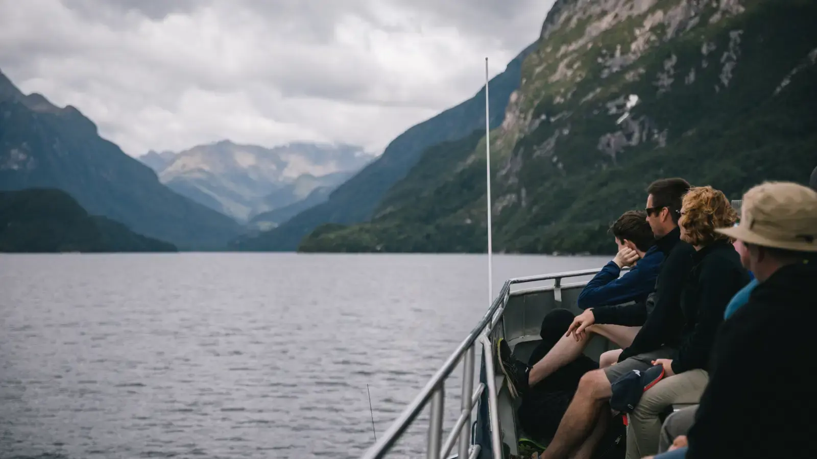 A group of people sit on the top deck of a modern catamaran as it cruises across Lake Te Anau.