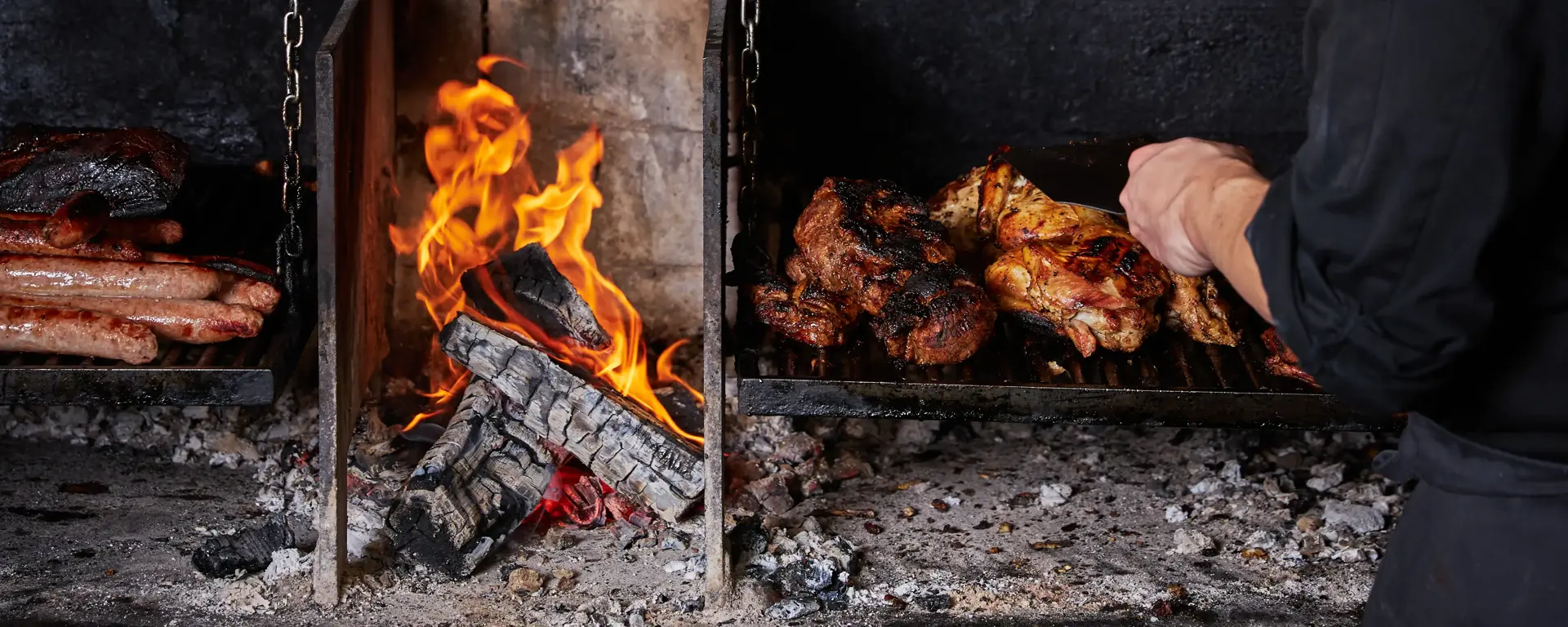 A chef standing at an indoor open fire BBQ tending to an assortment of meats. 