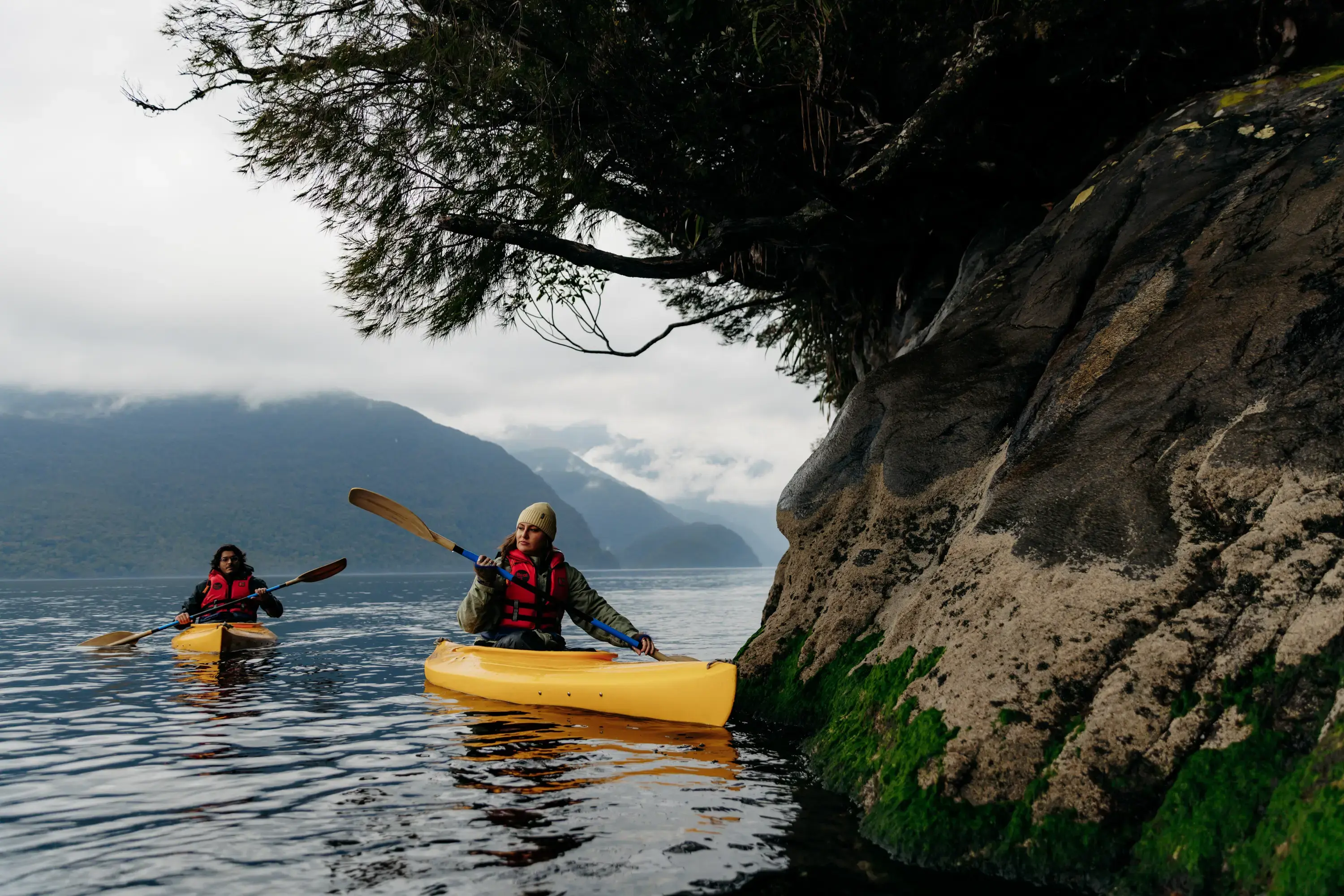 Two people kayaking beside a rocky, forested shoreline in Fiordland.
