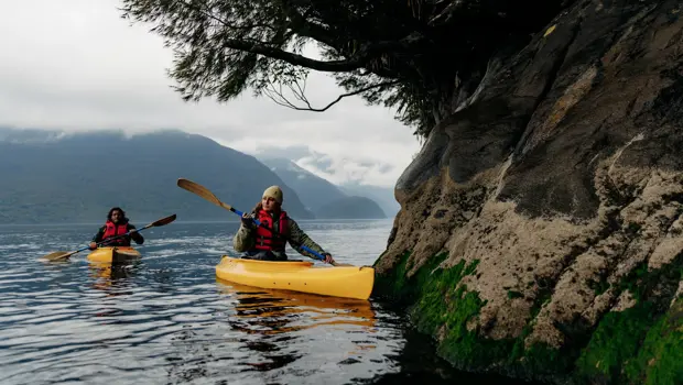 Two people kayaking beside a rocky, forested shoreline in Fiordland.