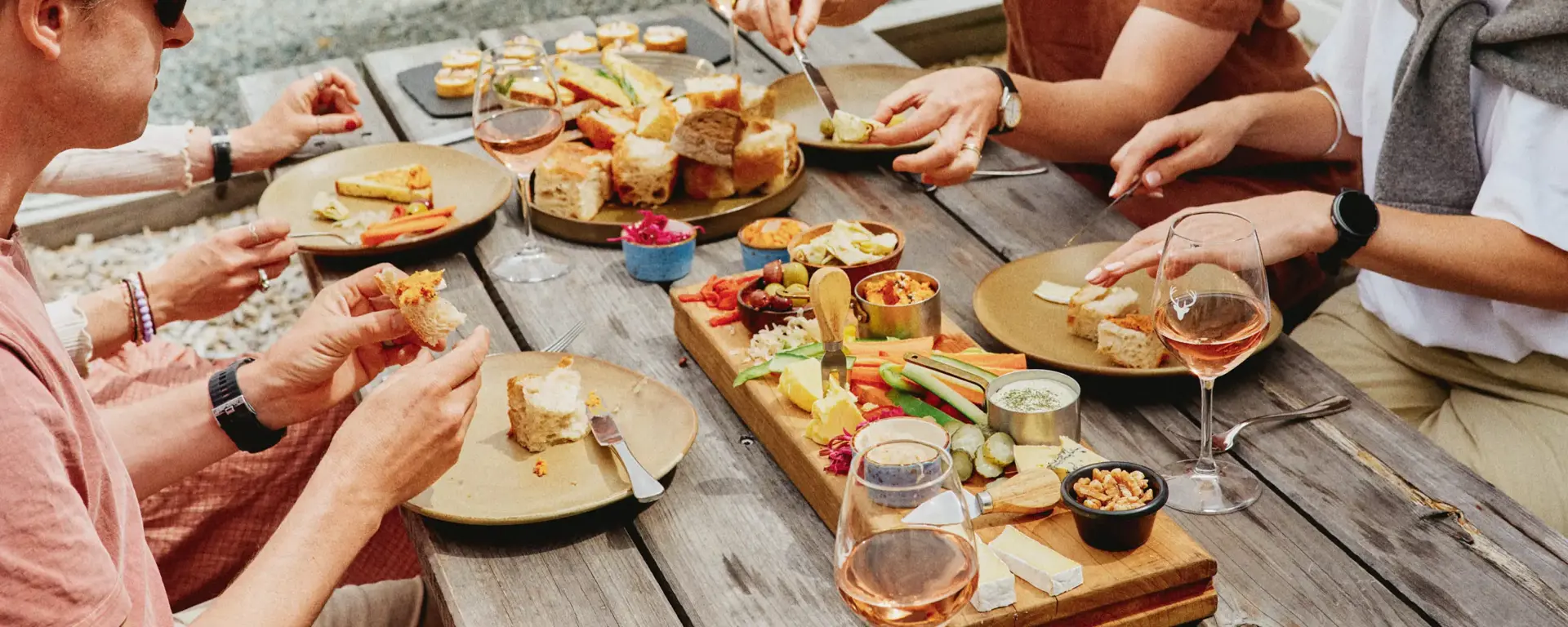 A group of friends enjoy shared platters of food and glasses of wine outside surrounded by green rolling hills.