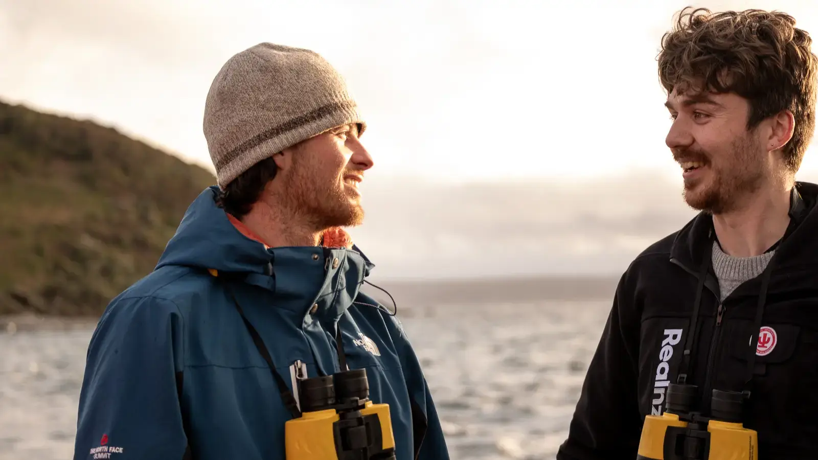 Two smiling guides in outdoor jackets, holding binoculars, chatting on a boat with the sea in the background.