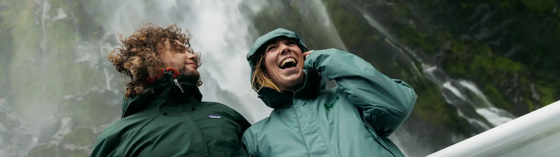 Two smiling guests on the outside deck of a cruise boat standing under a waterfall dressed in waterproof clothing.
