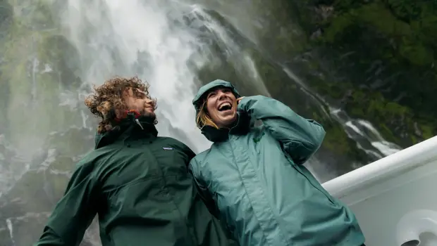 Two smiling guests on the outside deck of a cruise boat standing under a waterfall dressed in waterproof clothing.