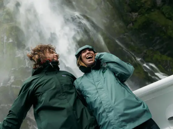 Two smiling guests on the outside deck of a cruise boat standing under a waterfall dressed in waterproof clothing.