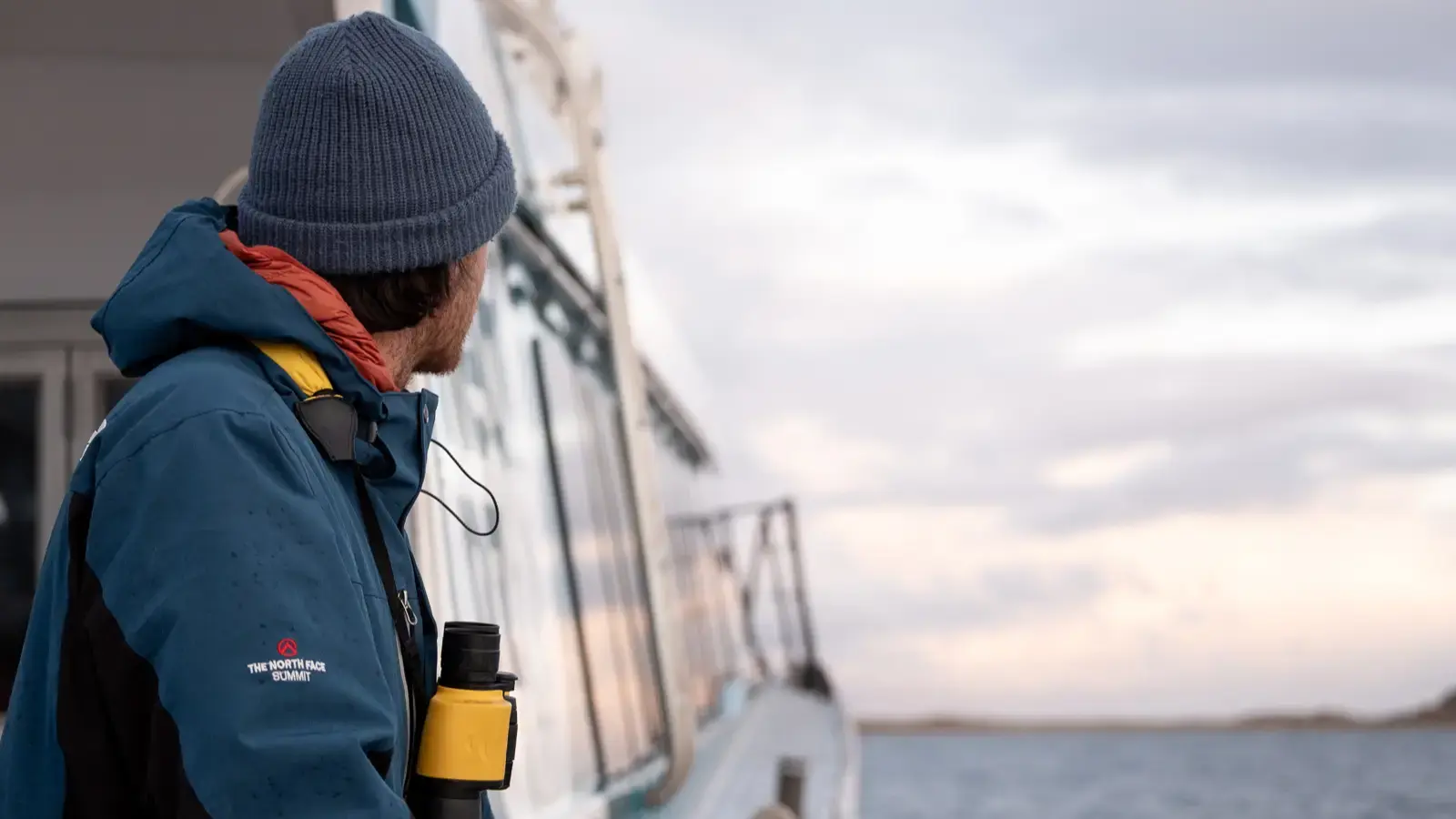 A man standing outside on a modern catamaran while it cruises across calm waters looks out ahead. 